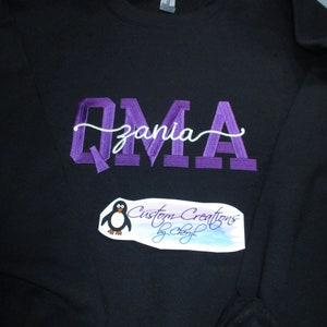 May include: A black sweatshirt with purple embroidered lettering that says "Qmzia" in a cursive font. The sweatshirt also has a small white penguin logo with the text "Custom Creations by Cheryl" underneath.