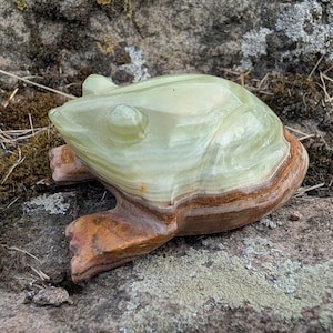 May include: A carved stone frog figurine featuring a smooth, pale green upper body with translucent qualities and a distinct, textured brown base. The sculpture displays natural banding and color variations throughout the polished mineral surface.