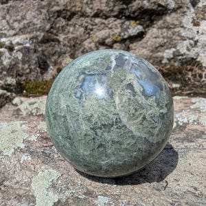 Pode incluir: Uma pedra esférica polida com uma textura mosqueada em tons de verde sálvia e musgo, apresentando inclusões brancas translúcidas. A superfície exibe padrões minerais naturais e variações de cor, criando uma aparência lisa e arredondada.