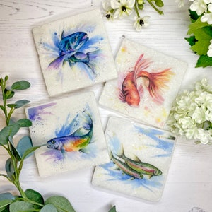 May include: Set of four square coasters featuring watercolor paintings of fish. Each coaster showcases a different fish species, including a blue trout, a goldfish, and two rainbow trout, with colorful splashes of paint.