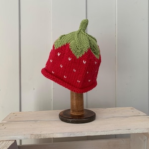 May include: A red and green knitted strawberry hat with white stitching. The hat is on a wooden display stand.
