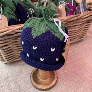 May include: A knitted strawberry hat with a green top and blue body. The hat has white stitching for the seeds and is displayed on a wooden stand.