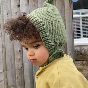 May include: A light green knitted hat with a pointed top, covering the head and ears. The hat has a textured design and a small green button detail. The child is wearing a yellow hooded sweatshirt.