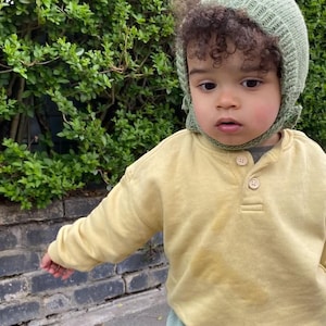 Puede incluir: Un niño con un gorro de punto verde claro y una camisa amarilla de manga larga con tres botones. El niño está de pie frente a un arbusto verde y una pared de ladrillos.