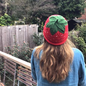 Op de afbeelding: Een rood en groene gebreide muts in de vorm van een aardbei. De muts heeft een groene, bladerige bovenkant en witte zaadjes op het rode lichaam.