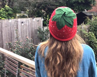 May include: A red and green knitted hat shaped like a strawberry. The hat has a green leafy top and white seeds on the red body.