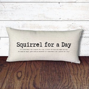 May include: A white pillow with black text that reads "Squirrel for a Day" and a quote about being a squirrel for a day. The pillow is on a wooden surface.