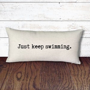 May include: A white rectangular pillow with the text "Just keep swimming." printed in black.