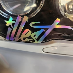 May include: Holographic "illest" sticker with a rainbow sheen, applied to a car headlight. The sticker's design features stylized lettering and a star-like element. The sticker is on a white car.