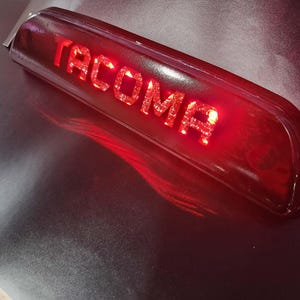 May include: A red, rectangular automotive light with the word "TACOMA" illuminated in bright red. The light has a curved design and is likely a third brake light for a vehicle.