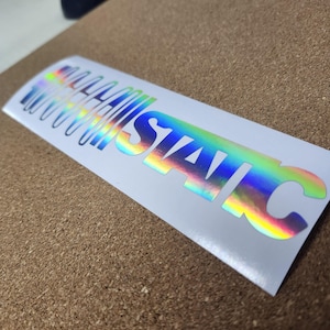 May include: A holographic sticker with the word "STATIC" in a bold, stylized font. The text has a rainbow gradient effect, with colors shifting across the letters. The sticker is rectangular and has a white background.
