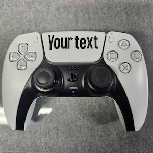 May include: A white and black game controller with the text "Your text" printed on the top. The controller has a black center section with two joysticks and a PlayStation logo. The buttons are white with black symbols.