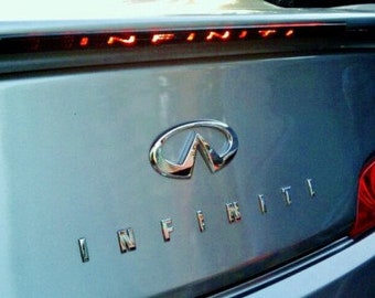May include: A silver Infiniti G35 car with the Infiniti logo and the word "INFINITI" written on the back. The car has a red brake light on the back.