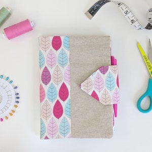 Puede incluir: Un cuaderno de tela con estampado rosa y azul, con una cubierta de lino y un bolsillo con solapa. El cuaderno tiene un bolígrafo rosa y unas tijeras amarillas a su lado.