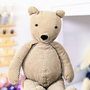 May include: A beige corduroy teddy bear with black button eyes. The bear is sitting with its legs crossed and its arms at its sides.