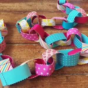May include: A colourful paper chain made of fabric scraps in pink, blue, yellow, and white. The chain is linked together in a continuous loop.