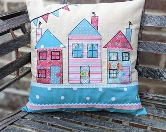 May include: A decorative pillow with a whimsical design of three houses with pink, blue, and white accents. The houses have small windows and doors, and the pillow has a blue and white polka dot trim.
