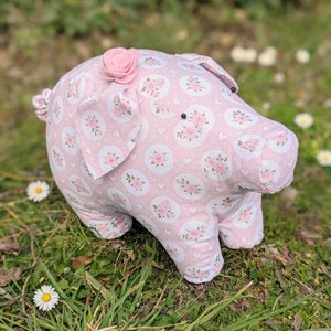 May include: A pink fabric pig with a floral pattern and a pink flower on its head. The pig is sitting on a bed of green grass.