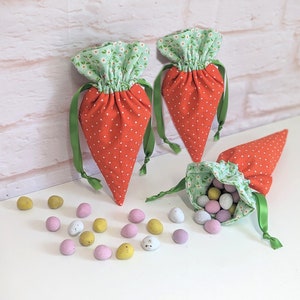 Può includere: Tre sacchetti di tessuto a forma di carota verde e bianca con pois bianchi. I sacchetti sono pieni di uova di caramelle color pastello.