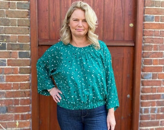 May include: A woman wearing a green and white patterned blouse with a gathered elastic waistband. The blouse has long sleeves and a round neckline. She is wearing blue jeans.