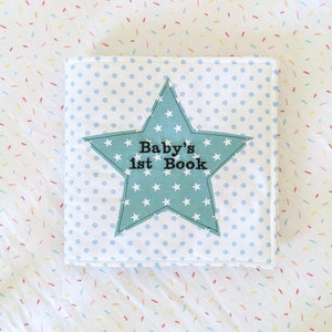 Può includere: Un libro per bambini blu e bianco con una copertina a forma di stella. La stella ha stelle bianche e il testo "Baby's 1st Book" in nero.