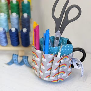 May include: A grey mug with a rainbow and cloud pattern fabric cover. The mug has four pockets for holding pens, pencils, scissors, and other small items. A pair of scissors, three pens, and a needle are in the pockets.
