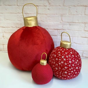 May include: Three red plush Christmas ornaments with gold trim. The largest ornament is plain red, the middle ornament has a white snowflake pattern, and the smallest ornament is plain red.