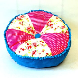 May include: Round decorative cushion with a patchwork design. The cushion features alternating segments of pink polka dots and floral patterns with red roses. It has a blue corduroy base and is trimmed with blue pom-poms.