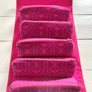 May include: A pink fabric organiser with five zippered pouches. Each pouch has a geometric pattern with pink and purple tones.