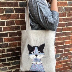 May include: A beige linen tote bag with a black and white cat face appliqué. The cat has a blue and white floral pattern on its body and a white lace collar.