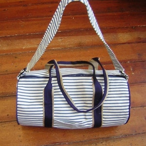 Könnte beinhalten: Eine weiße und blaue gestreifte Reisetasche mit einem marineblauen Trageriemen und Besatz. Die Tasche hat einen Reißverschluss und zwei obere Tragegriffe.