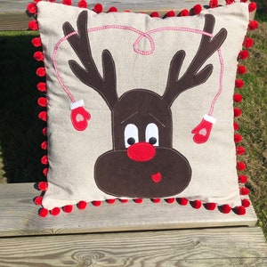 May include: A beige cushion with a brown reindeer face and red pom-poms around the edge. The reindeer has a red nose and white eyes.