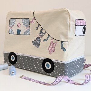 May include: A cream-coloured sewing machine cover with a grey and white polka dot trim. The cover features a whimsical design of a camper van with clothes hanging on a line. The camper van has a blue window with a pink flower in a pot. The cover is sitting on a white surface with a pink measuring tape.
