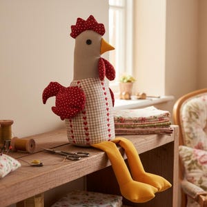 May include: A fabric rooster doll with a red comb, wattle, and polka-dot wings. The body is checkered with heart accents. It has yellow legs and sits on a wooden shelf with sewing supplies. A floral pin cushion and spools of thread are also visible.