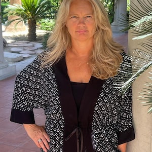 May include: A black and white patterned kimono-style jacket with three-quarter sleeves. The jacket has a black collar and a tie closure. The woman is wearing a black top underneath.