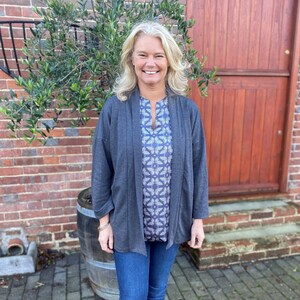 May include: A woman wearing a dark gray cardigan over a white and gray patterned blouse and blue jeans. She is standing in front of a wooden door.