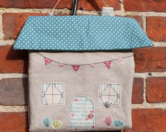 May include: A beige fabric house-shaped wall hanging with a blue and white polka dot roof. The house has a small window, a door, and a string of pink and white flags. The text "is. Ho where th home is wh where the is. Ho re the ome is whe where the is. Ha" is printed on the house.