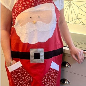 May include: Red Christmas apron with a Santa Claus design. The apron has a white beard, black eyes, and a black belt with a silver buckle. The apron has two red mitten pockets with white snowflake designs.