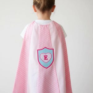 May include: A pink and white superhero cape featuring alternating solid pink and white polka-dot panels. The centre of the cape displays a light blue shield-shaped emblem with a pink border, containing a white heart with a pink letter E inside.