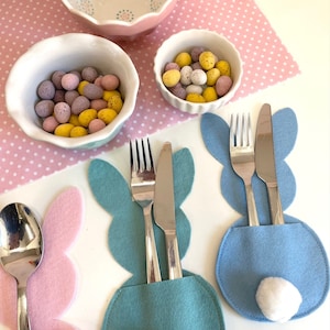 May include: Three Easter bunny-shaped felt place card holders. One is pink with a spoon, one is blue with a fork and knife, and one is teal with a fork and knife. Each holder has a white cotton ball for a tail.