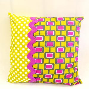 Può includere: Un cuscino giallo con un motivo geometrico rosa e grigio su un lato e un motivo a pois bianchi sull'altro lato. Il cuscino ha un bordo rosa a festoni.