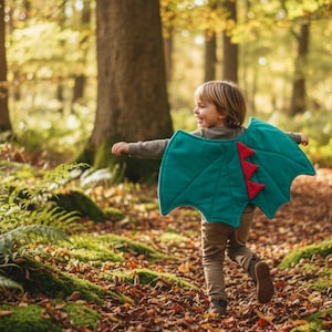 May include: A child wearing a teal dragon wing costume with red accents, running through a forest. The wings are attached to a gray shirt. The background features trees, fallen leaves, and green moss.