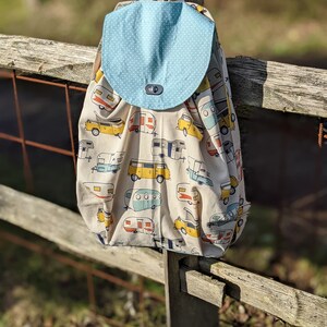 May include: A beige backpack with a light blue polka dot flap. The main body of the bag is patterned with colourful camper illustrations. The bag is hanging on a wooden fence. The bag is suitable for carrying items.