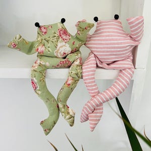May include: Two plush frog dolls. One frog is green with a floral pattern of pink and white roses. The other frog is pink and white striped. Both frogs have black bead eyes and are sitting on a white shelf.