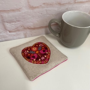 May include: A heart-shaped coaster with a pink, red, and orange floral design and two birds. The coaster is made of a beige linen fabric with a pink border.