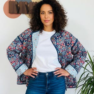 May include: A navy blue floral print jacket with pink and orange flowers. The jacket is worn over a white t-shirt and blue jeans. The jacket has a light blue lining and button closure. The background includes a wall decoration and a green plant.