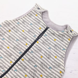 May include: A gray and white striped baby sleep sack with a zipper closure. The fabric features a repeating pattern of clouds with stars and crescent moons.