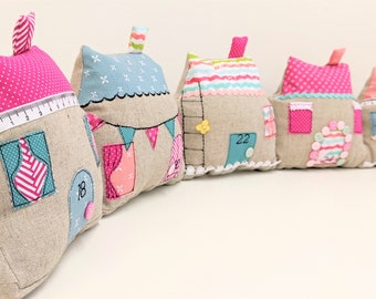 May include: Five small, fabric houses in a row. Each house is made of beige linen with different colored fabric accents. The houses are decorated with buttons, embroidery, and numbers. The houses are arranged in a row on a white surface.