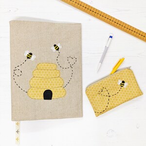 May include: A beige linen pencil case with a beehive and bees embroidered on it. A smaller matching pencil case with a beehive pattern is also shown.