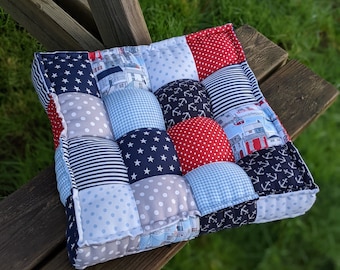 May include: A blue and red patchwork cushion with a nautical theme. The cushion is made of different fabrics with various patterns, including stars, anchors, stripes, and polka dots.